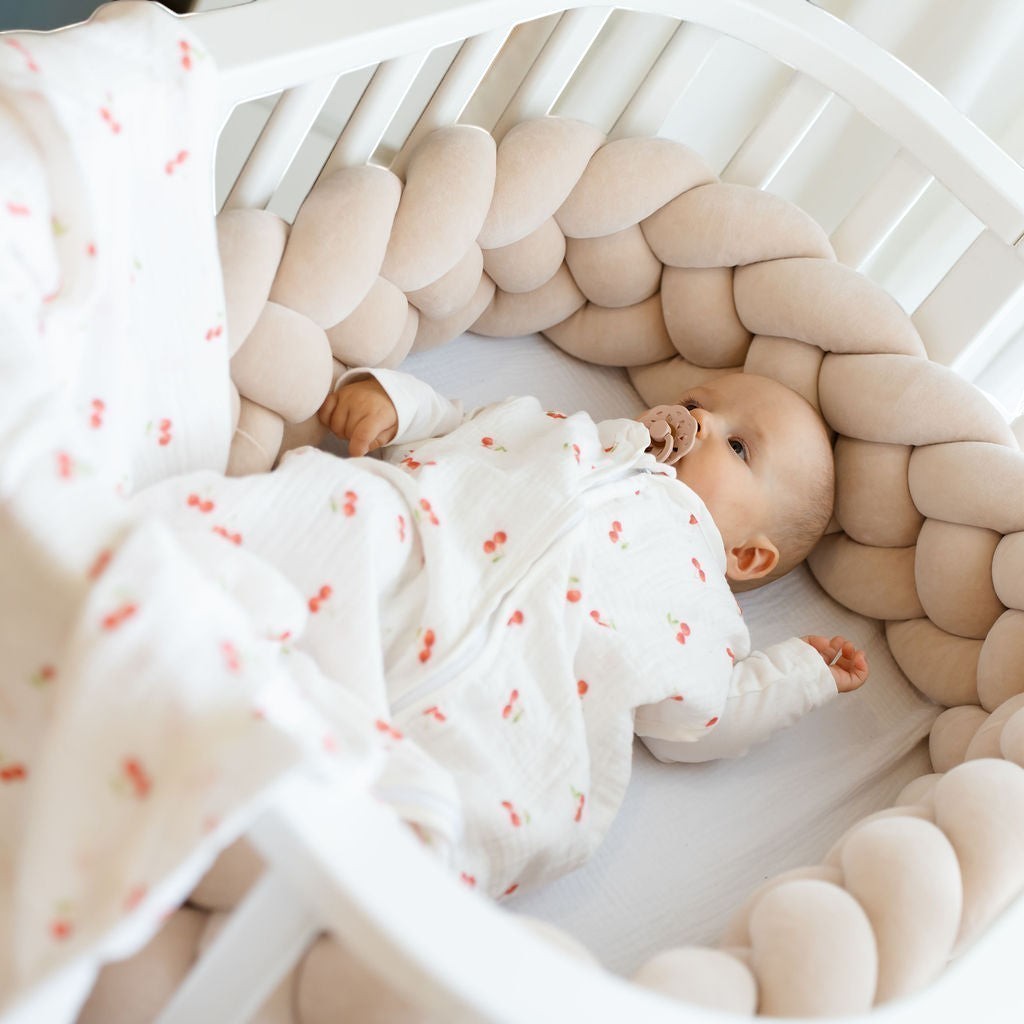 Baby Natur white cherry print sleeping bag with a beige braided crib bumper and a baby wearing the sleeping bag.