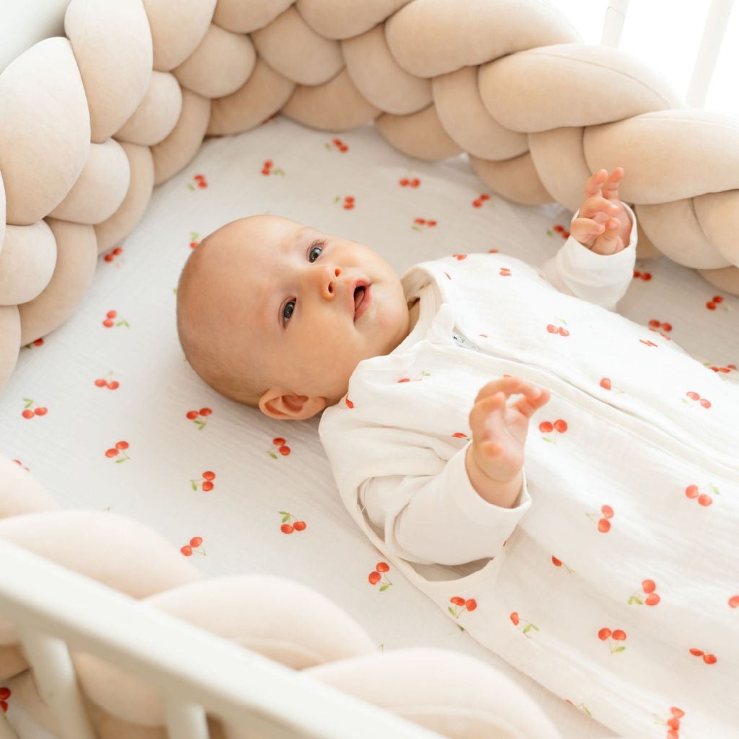 Baby Natur white cotton crib sheet with a red cherry print, featuring a baby lying on it.