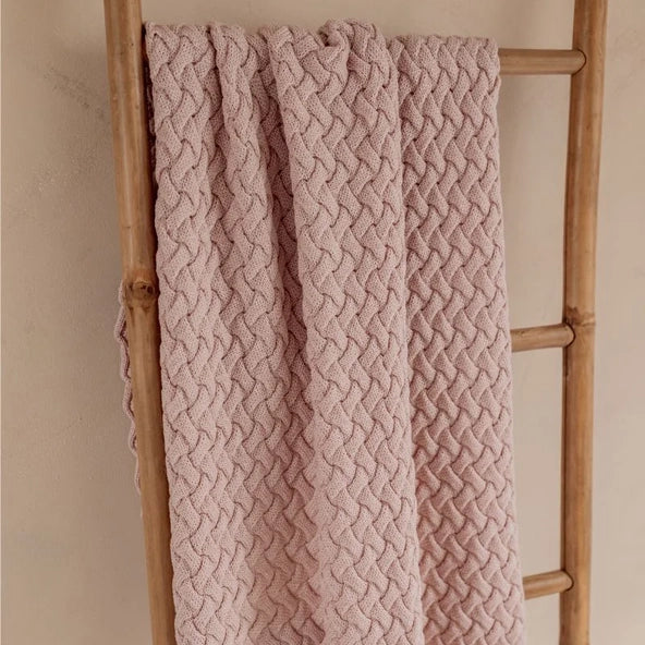 A dusty pink cable-knit blanket draped over a rustic wooden ladder against a neutral beige wall.